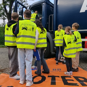 Eine Gruppe von Kindern steht vor einem LKW und trägt neongelbe Warnwesten mit zwei waagerechten, grauen Reflexionsstreifen auf der Rückseite.