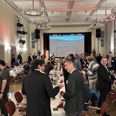 Männer und Frauen in einem feierlichen Festsaal. Im Hintergrund eine Bühne mit Leinwand.