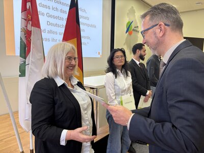 Ein Mann mittleren Alters im Anzug mit Brille (rechts im Bild) überreicht einer Frau mit weißblonden, schulterlangen Haaren und Brille ein Dokument. 