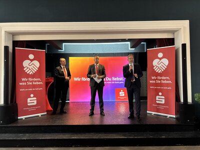 Drei Männer mittleren Alters in Anzügen stehen auf einer Bühne. Der Mann rechts im Bild hält in der rechten Hand ein Mikrofon. 