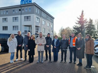 Eine Gruppe von Menschen steht vor einem weiß-grauen Haus mit vielen Fenstern. Auf dem Dach steht in blauer Schrift MRA. 