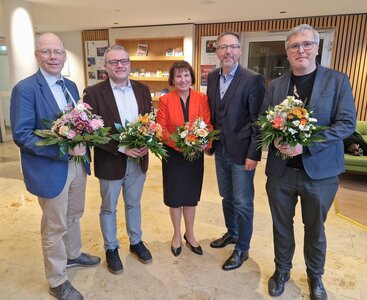 Vier Männer und eine Frau stehen mit Blumensträußen in den Händen in einem beleuchteten Foyer und lächeln in die Kamera. 