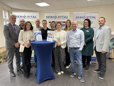 Männer und Frauen stehen in Zweierreihe vor drei Hängebannern mit dem Aufdruck SENIO-VITAL vor einem blauen Stehtisch.