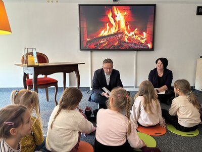 Ein Mann mit einem aufgeklappten Buch in der Hand sitzt vor einem Monitor mit loderndem Kaminholz. Im Vordergrund mit dem Rücken zur Kamera sitzen kleine Mädchen Grundschulalter.