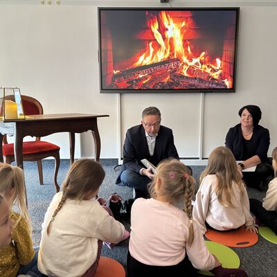 Ein Mann mit einem aufgeklappten Buch in der Hand sitzt vor einem Monitor mit loderndem Kaminholz. Im Vordergrund mit dem Rücken zur Kamera sitzen kleine Mädchen Grundschulalter.