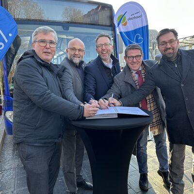 F&uuml;nf M&auml;nner mittleren Alters stehen um einen schwarzen Stehtisch mit Stiften in den H&auml;nden. Im Hintergrund ein Verkehrsbus und zwei Beachflaggen mit dem Logo 1plus7 und vom Landkreis Oberhavel. 