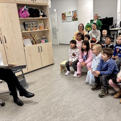 Eine Frau mit Arztkittel (links im Bild) sitzt auf einem Hocker und liest einer Gruppe von Vorschulkindern aus einem Buch vor.