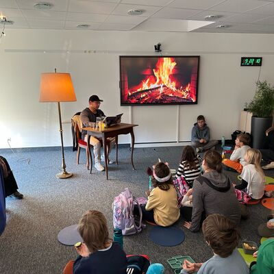 Eine Gruppe von Kindern sitzt auf dem Boden mit dem Rücken zur Kamera. An der Wand hängt ein Monitor mit Kaminfeuerbild. An einem Tisch sitzt ein Mann mit Brille und Basecap, der aus einem Buch liest.