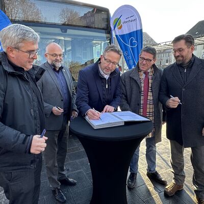 Fünf Männer mittleren Alters stehen um einen schwarzen Stehtisch. Im Hintergrund ein Verkehrsbus und zwei Beachflaggen mit dem Logo 1plus7 und vom Landkreis Oberhavel.