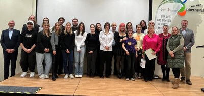 Eine Gruppe von Männer und Frauen stehen auf einer Bühne und schauen in die Kamera. 