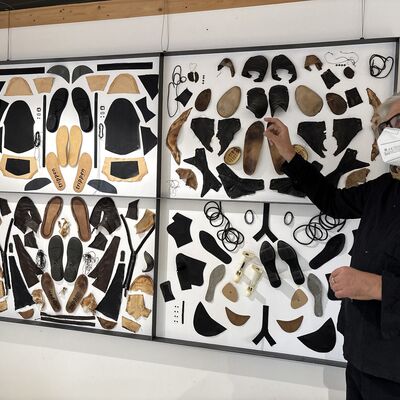 Ein Mann mit schwarzem Pullover, schwarzer Brille und einer FFP2-Maske vor dem Mund steht vor einer Wand mit Bilderrahmen, in denen Schuhsohle hinter Glas ausgestellt sind.