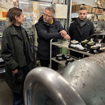Zwei M&auml;nner und eine Frau stehen in einer Fabrikhalle, in der Schuhe hergestellt werden.