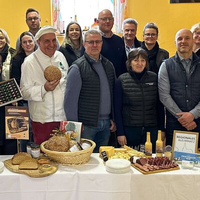 Eine Gruppe von M&auml;nnern und Frauen stehen vor einem mit Nahrungsmitteln gedeckten Tisch und schauen in die Kamera.
