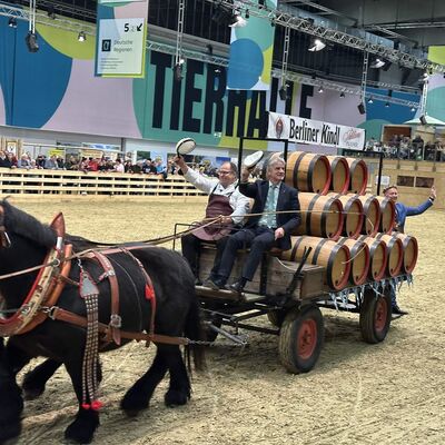 Zwei &auml;ltere M&auml;nner sitzen in einer landwirtschaftlichen Pferdekutsche, die mit F&auml;ssern aus Holz beladen ist und von zwei dunklen Pferden durch eine Arena gezogen wird.