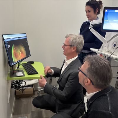 Zwei M&auml;nner hocken vor einem Monitor. Sie schauen auf eine mikroskopische Aufnahme vom inneren des Auges. Eine junge Frau steht hinten rechts im Bild.