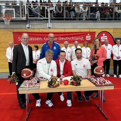 F&uuml;nf M&auml;nner und eine Frau sitzen und stehen um einen Tisch in einer Turnhalle. Der Tisch steht auf einem roten Teppich. Im Hintergrund steht eine Beachflag und an der Wand h&auml;ngt ein Banner mit dem Sparkassen-Logo.