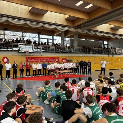 Kinder und Jugendliche sowie Erwachsene sitzen und stehen auf dem Boden einer Turnhalle. Im Hintergrund h&auml;ngt an einer Wand das Logo der Mittelbrandenburgischen Sparkasse.