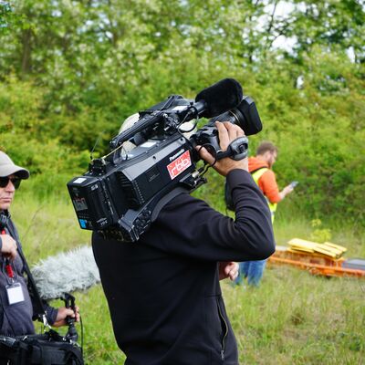 Medienvertreter des regionalen Fernsehsenders RBB mit Kameramann und Assistenten, stehend am Gleis der Zugstrecke im &Uuml;bungsabschnitt MANV. Im Hintergrund ein Beobachter bei Sichtung von Rettungsmaterial.