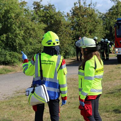 Zwei Personen in Rettungskr&auml;fteuniformen stehen im Bildvordergrund auf einer gr&uuml;nen Rasenfl&auml;che. Im Hintergrund steht ein rotes Feuerwehrauto und weitere Rettungskr&auml;fte.