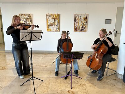 Zwei M&auml;dchen im Teenageralter und eine Frau musizieren gemeinsam. Im Hintergrund h&auml;ngen an der Wand hochformatige Bilder mit Naturmaterialien gefertigt. 