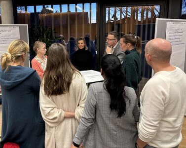 Eine Gruppe von mehreren Frauen und zwei M&auml;nnern steht um einen Stehtisch. Im Hintergrund stehen Pinnw&auml;nde mit A3 Bl&auml;ttern. 