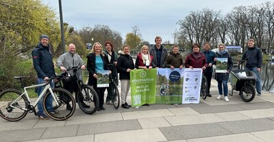 M&auml;nner und Frauen mit Fahrr&auml;dern und dem Stadtradeln-Banner 2026stehen in einer Reihe an einem Fluss, vermutlich der Havel, und posieren f&uuml;rs Pressefoto. 