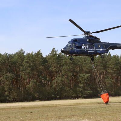 Ein blauer Hubschrauber schwebt wenige Meter &uuml;ber einem bewaldeten Boden. Er transportiert einen L&ouml;schwassereimer.