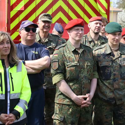 Eine Frau mit neongelber Jacke und M&auml;nner in Uniform der Bundeswehr stehen vor einem Einsatzfahrzeug des Brandenburgischen Katastrophenschutzes.