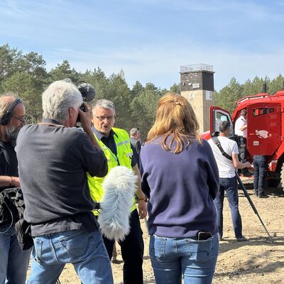Ein &auml;lterer Mann mit Brille und neongelber Warnweste steht im Kreis umringt von M&auml;nnern und einer Frau mit Kameras und Mikrofonen . Im Hintergrund sieht man ein gro&szlig;es, rotes Spezialfahrzeug des Katastrophenschutzes Brandenburg.