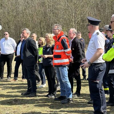 M&auml;nner und Frauen in Uniformen und Einsatzwesten sowie Kameraleute stehen seitlich von der Kamera abgewendet auf einer Wiese. Im Hintergrund sind B&auml;ume zu erkennen.