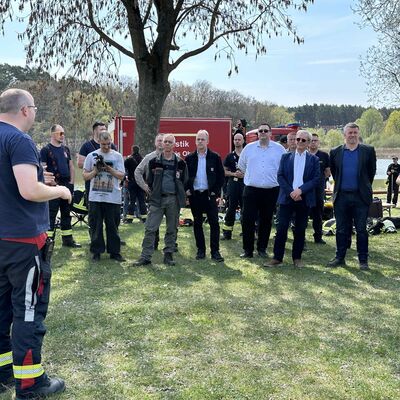 M&auml;nner in Einsatzbekleidung des Katastrophen- und Brandschutzes stehen auf einer Wiese vor einem roten Einsatzfahrzeug des Katastrophen- und Brandschutzes. Im Hintergrund B&auml;ume, Wald und der Beetzer See.
