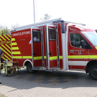 Ein rotes Einsatzleitungsfahrzeug der Feuerwehr parkt auf einem gepflasterten Steinboden bei ge&ouml;ffneten T&uuml;ren. Zwei M&auml;nner in Einsatzkleidung stehen rechts und links vor dem Fahrzeug.