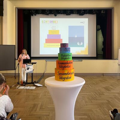 Turm der Medienm&uuml;ndigkeit des Pr&auml;ventionsprogramms ECHT DABEI auf einem wei&szlig;en Stehtisch, im Hintergrund spricht ECHT-Coach Sabrina Sebbesse