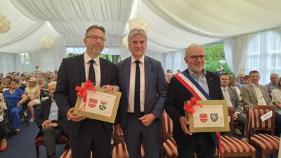 Drei M&auml;nner mittleren Alters mit Anz&uuml;gen stehen in einem Festzelt und schauen direkt in die Kamera. Der Mann rechts im Bild tr&auml;gt eine Sch&auml;rpe quer &uuml;ber die Schulter und h&auml;lt wie der Mann links im Bild einen Geschenkgutschein in den H&auml;nden. 