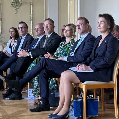 Vier M&auml;nner und drei Frauen sitzen in einer Stuhlreihe in einem festlichen Saal.