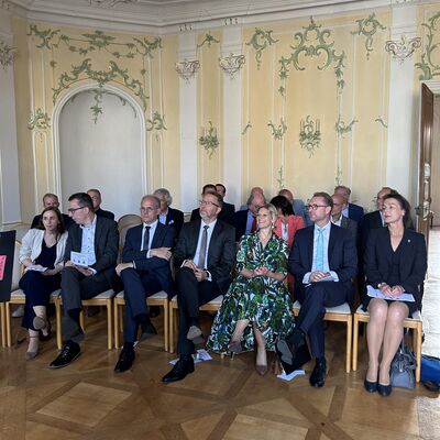 Vier M&auml;nner und drei Frauen sitzen in einer Stuhlreihe in einem festlichen Saal.