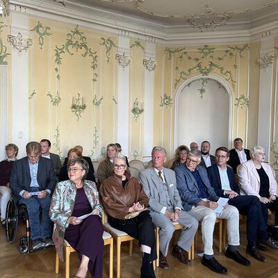 Drei M&auml;nner und drei Frauen sitzen in einer Stuhlreihe in einem festlichen Saal.