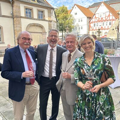 Drei M&auml;nner und eine Frau in festlicher Kleidung stehen auf einem Marktplatz mit Gl&auml;sern in den H&auml;nden und lachen in die Kamera.