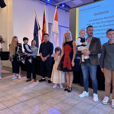 M&auml;nner, Frauen und Kinder vor einer Leinwand in einem Festsaal anl&auml;sslich der aktuellen Einb&uuml;rgerungsveranstaltung in der KulturFuge.