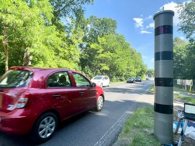 Eine Bundesstraße mit mehren Autos. Im Vordergrund ein dunkelroter Kleinwagen, der an einer Blitzersäule vorbeifährt.
