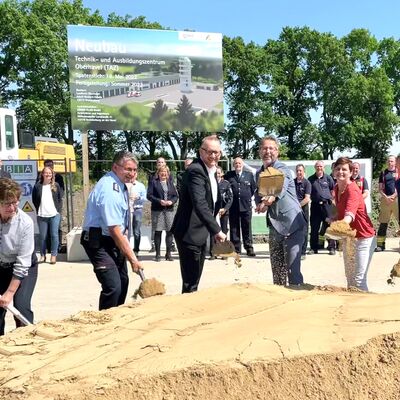 Im Vordergrund des Bildes ist ein Sandhaufen. Dahinter stehen von links nach rechts Andrea Will, Gerd Ritter, Matthias Rink, Alexander Tnnies und Maja Dorfmeister, die mit einem Spaten Sand in die Luft schippen. Im Hintergrund ist das Baustellenschild zu sehen.