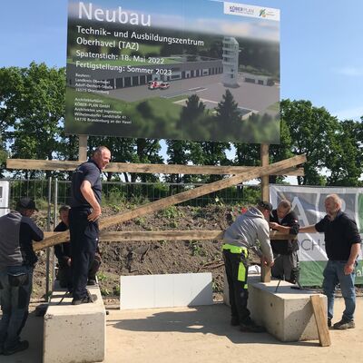 Vier Männer in schwarzer Arbeitskleidung stellen das Baustellschild auf.