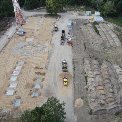 Blick von oben auf das Baulfeld. In der linken Bildhälft sind zehn Fundamente für die Säulen zu sehen. In der rechten Bildhälfte sind mehrer Sandhaufen nebeneinander angelegt. Zwischen den beiden Bereichen ist eine Baustraße mit mehreren Baufahrzeugen.