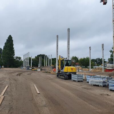 Blick von der Straße auf die Säulen des Rohbaus. Im rechten Bildrand steht ein gelber Minibagger, davor Paletten mit Baumaterialien.