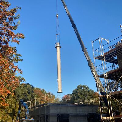 Blauer Himmel im Hintergrund, rechts im Vordergrund Teile eines Baugerstes, dahinter erstreckt sich ein Kranarm nach links oben. An den Ketten des Kranarms hngt der Schornstein fr das Heizhaus. Er schwebt etwas ber den Rohbau, der unten mittig im Bild zu erkennen ist.